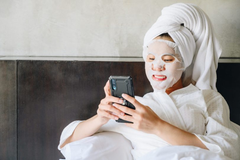 happiness-young-woman-sitting-on-bed-and-using-smart-phone-while-applying-facial-mask-for-enhance-her-skin