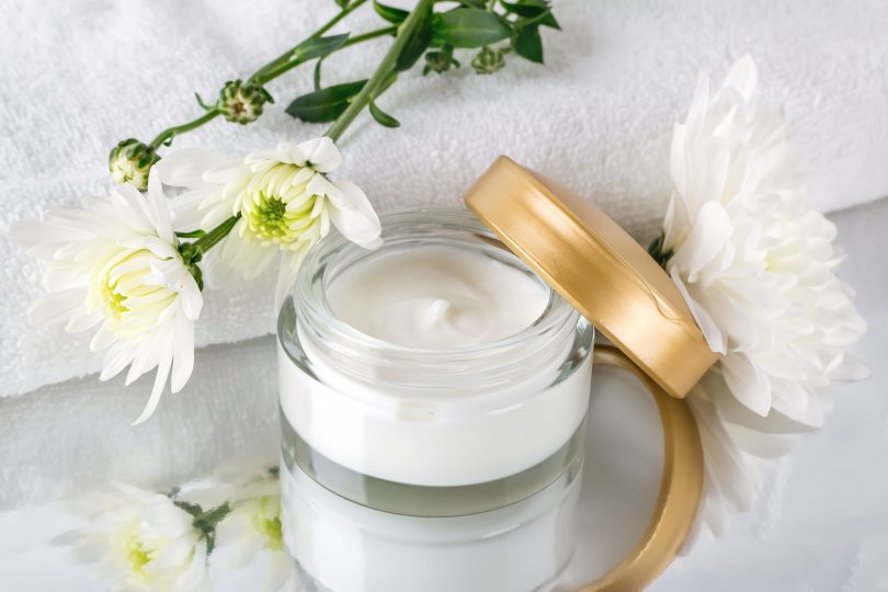 facial-cream-in-an-open-glass-jar-with-a-golden-lid-next-to-fresh-chrysanthemum-buds-and-a-white-terry-towel-on-a-mirror-surface-beauty-skincare-and-cosmetology
